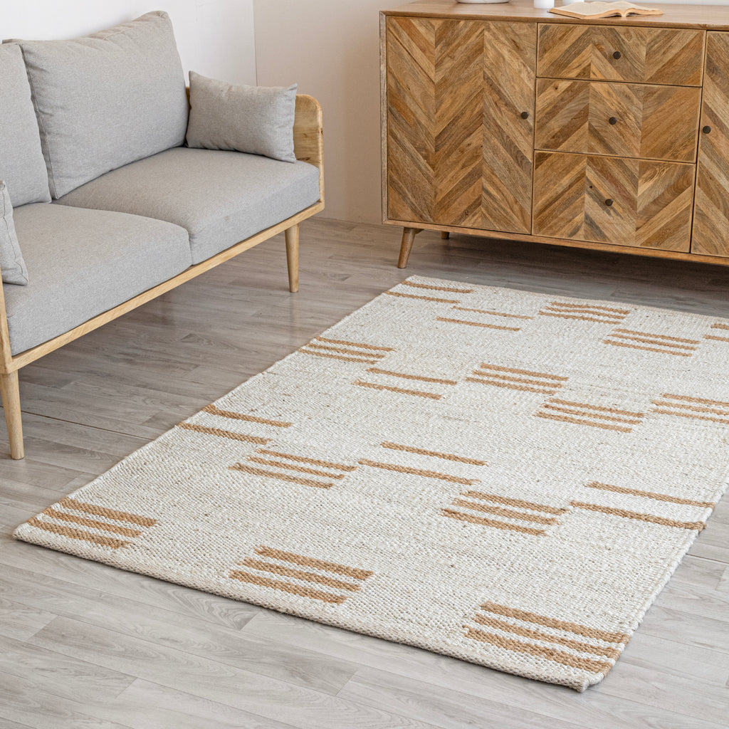 A striped natural jute rug with off white and beige colors, placed on a floor under a modern sofa and a wooden cabinet.