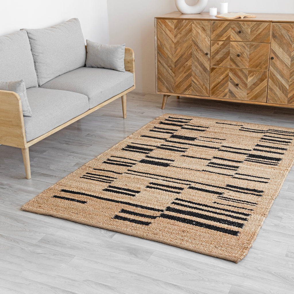 A striped natural jute rug in beige and black, laid out on a floor next to a gray sofa and a wooden TV stand, with natural wood walls and a white coffee table in the background.